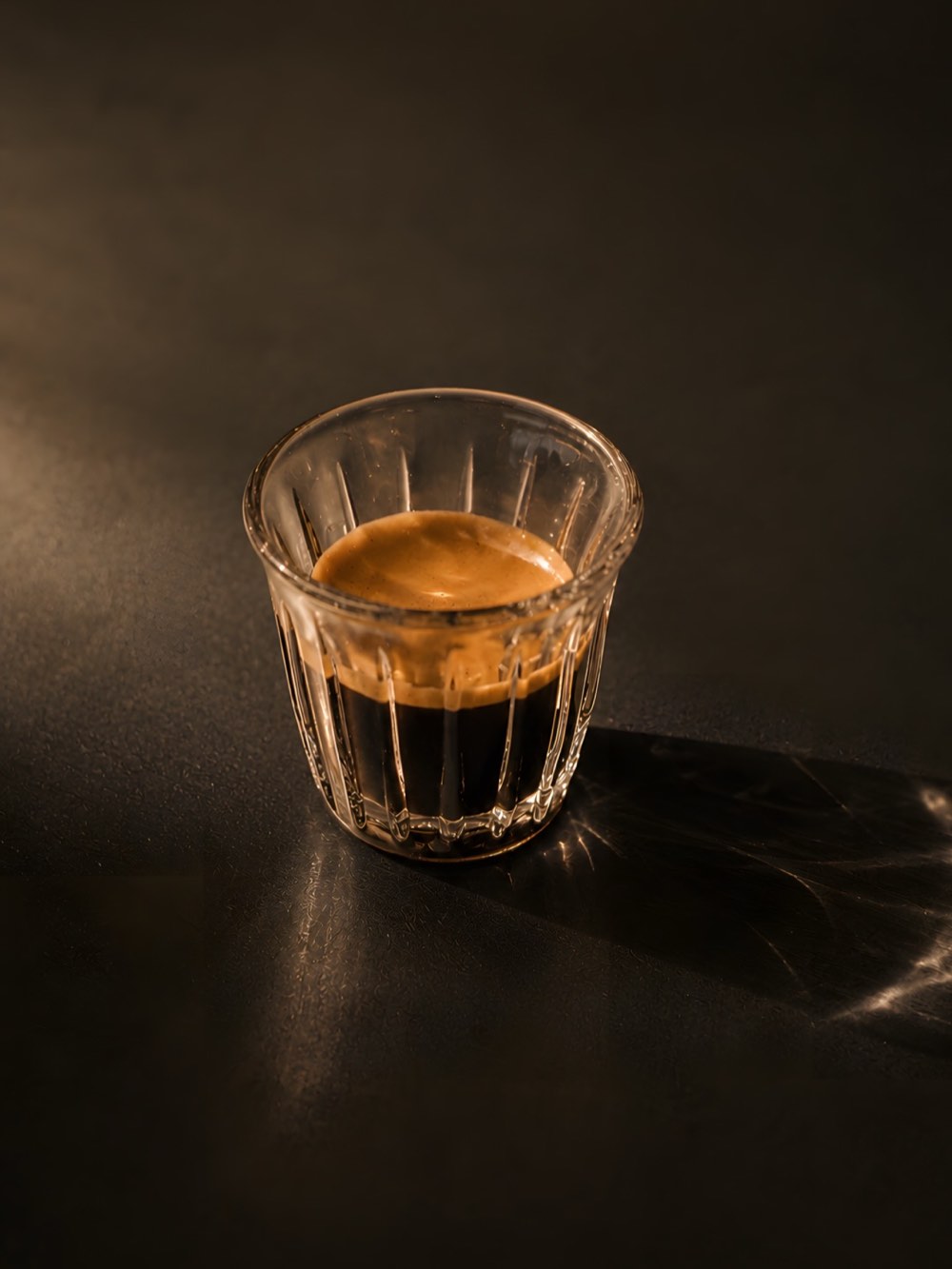 A single espresso in a ribbed glass, lit from one side on a black surface.