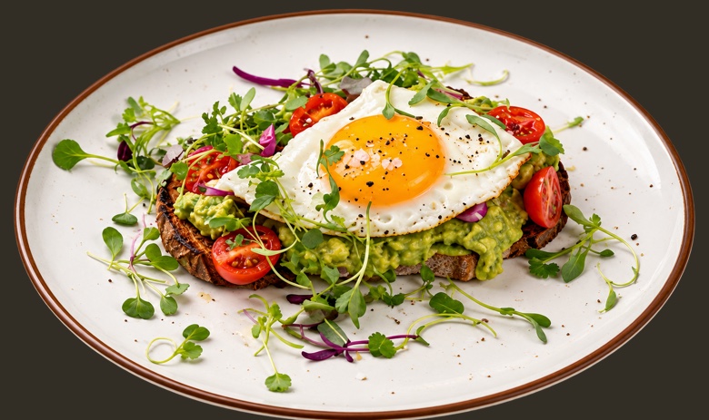 Avocado toast on a rimmed plate: mashed avocado over dark rye under a sunny side egg, with cherry tomatoes and microgreens.