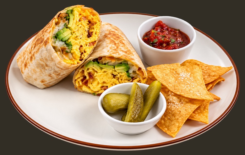 Breakfast burrito cut in half on a rimmed plate, showing scrambled egg, bacon and avocado, with salsa, pickles and tortilla chips alongside.