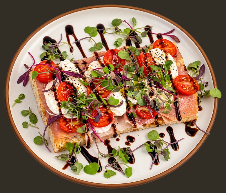 Burrata and prosciutto toast on a rimmed plate, with cherry tomatoes, microgreens and a balsamic drizzle.