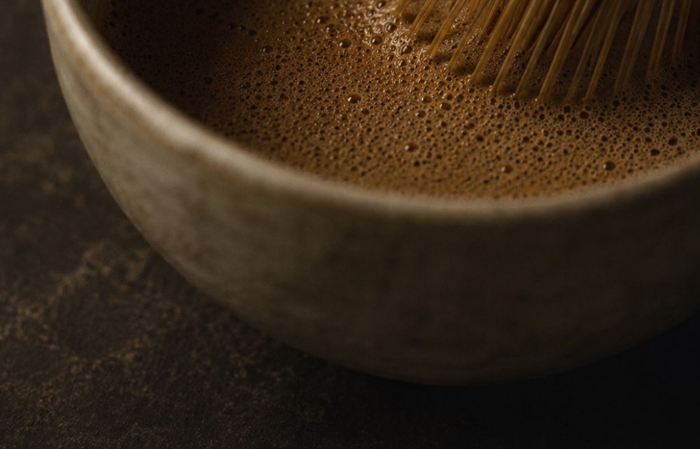 Hojicha whisked to a fine foam in a pale ceramic bowl, the tips of a bamboo chasen still resting in the cup.